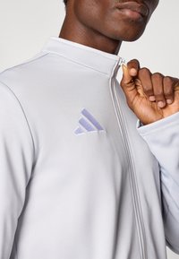 Ανοιχτό γκρι ζακέτα με φερμουάρ από μαλακό ύφασμα, με ψηλό γιακά και διακριτικό υφή επιφάνειας. Μωβ κεντητό λογότυπο στο αριστερό μέρος του στήθους.