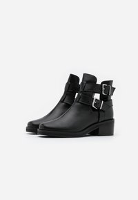 Bottines en cuir noir avec un nez légèrement pointu, comportant deux sangles ajustables et des boucles métalliques. Talon carré modéré.