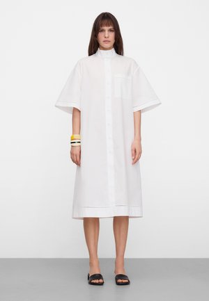 Kobieta ubrana w luźną białą sukienkę koszulową midi z krótkimi rękawami, zapinana z przodu na guziki, z kieszenią na piersi, czarne sandały na płaskim obcasie i kolorowe bransoletki na lewej ręce.