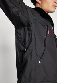 Veste légère noire avec une surface texturée, dotée de poches zippées et de tirettes de zip rouges pour une touche de détail.