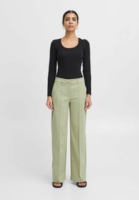 Črn bodi z dolgimi rokavi v kombinaciji z visokim pasom in širokimi svetlo zelenimi hlačami; zasnova je minimalistična in tekstura gladka.