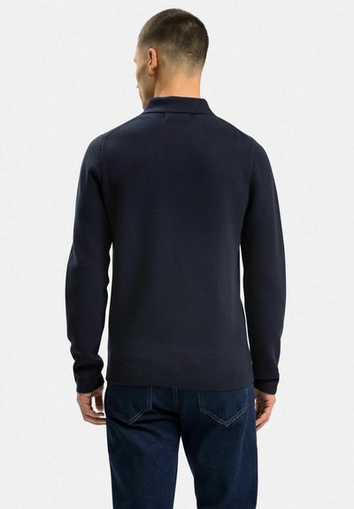Mörk marinblå långärmad polo med krage, slät textur och skräddarsydd design. Bärs tillsammans med blå denimjeans, vilket visar en enkel bakvy.