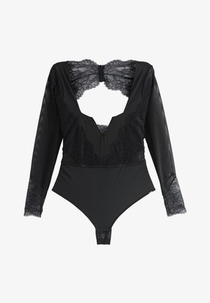 Femme portant un body en dentelle noire transparente à manches longues, posant à l'intérieur devant des rideaux beiges et un canapé blanc.