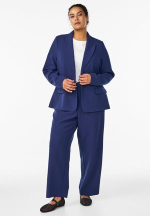 Kobieta z kołowymi kolczykami, ubrana w niebieski garnitur, biały top i czarne płaskie buty, stojąca na tle jednolitego białego tła.