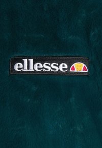 Turkusowy futerko z tkaniny z przyszytą czarną metką z białym napisem "ellesse" oraz kolorowym logo w czerwonych i żółtych półokręgach.
