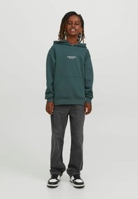 Felpa verde scuro realizzata in un tessuto morbido, con tasca frontale e dettaglio del logo bianco. Abbinata a jeans grigi larghi e sneaker nere.