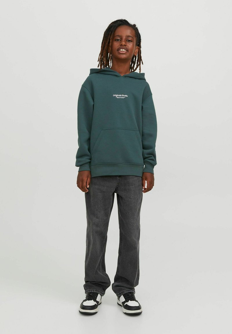 Felpa verde scuro realizzata in un tessuto morbido, con tasca frontale e dettaglio del logo bianco. Abbinata a jeans grigi larghi e sneaker nere.