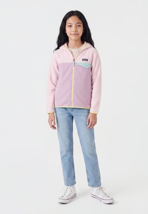 Fille aux longs cheveux noirs portant un sweat à capuche zippé rose et violet, un jean bleu clair et des baskets blanches, debout sur un fond blanc.