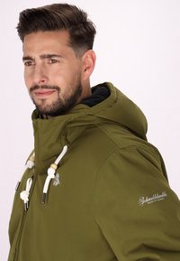 Giacca verde oliva con cappuccio, caratterizzata da coulisse bianche, dettagli in legno e un logo sulla manica con una texture liscia.