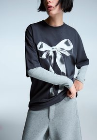 Sort oversized t-shirt med et sølvfarvet bue-grafik, parret med lysegrå lange ærmer og matchende grå sweatpants. Glat tekstur.