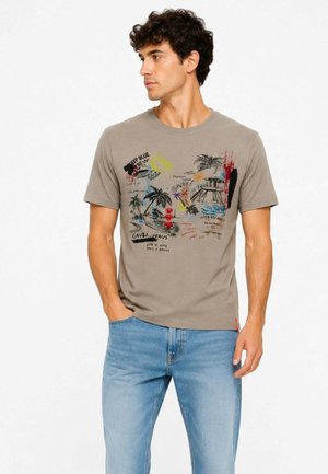 Uomo che indossa una t-shirt beige a maniche corte con grafiche tropicali colorate e testo, abbinata a jeans blu chiaro, guarda di lato.