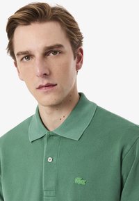 Vihreä polo-paita, jossa on teksturoitu kangas, klassinen kaulus, kolmen napin läppä ja vihreä brodeerattu logo rinnassa.