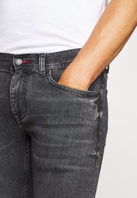 Jeans de denim negros de corte slim con un ligero desvanecimiento, diseño de cinco bolsillos y un acento rojo en el botón. El material tiene una textura suave.