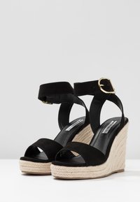 Black suede wedge sandals with an ankle strap, featuring a jute-wrapped platform, open toe, and gold-tone buckle detail.