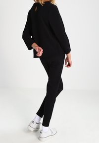 Czarny top z długim rękawem z wycięciem w kształcie otworu z tyłu, w połączeniu z czarnymi dopasowanymi spodniami i białymi trampkami, pokazany od tyłu.