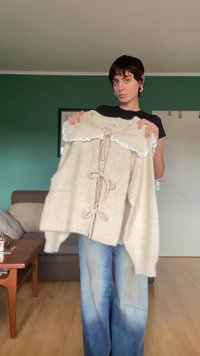 Suéter de malha bege com colarinho de renda, apresentando laços na frente e um corte solto. O tecido parece suave e texturizado.