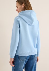 Sweat à capuche bleu clair en tissu doux, avec une capuche, des poignets côtelés et une coupe décontractée. Porté avec un jean bleu, affichant un style décontracté.