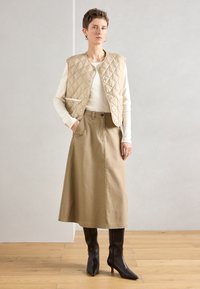 Colete acolchoado bege com fechos de pressão, saia comprida bege e botas castanhas de salto até ao joelho. Design simples e funcional, com poucos detalhes.