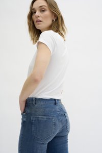 Bela kratka majica s kratkimi rokavi v kombinaciji z modrimi jeans hlačami z visokim pasom. Hlače imajo zadnje žepke in gladko, rahlo zbledelo teksturo.