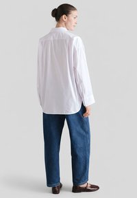 Biała koszula z długim rękawem i guzikami, zestawiona z luźnymi niebieskimi dżinsami i brązowymi balerinami, widziana od tyłu. Gładka tkanina i klasyczny krój.