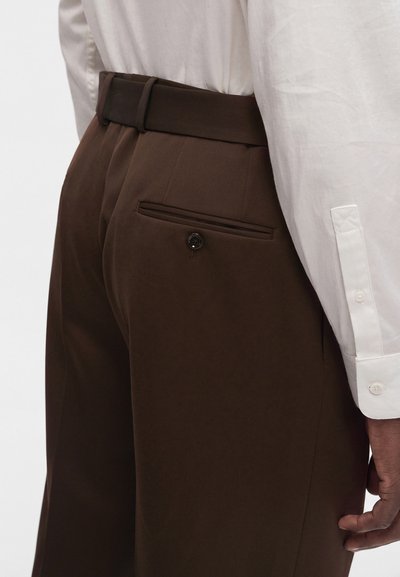 Gros plan d'une personne portant un pantalon marron sur mesure avec une poche arrière boutonnée et une chemise blanche à manches longues.