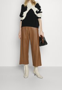 Pull à motif noir et crème, pantalon large en cuir marron, bottines beiges, tenant un sac noir avec une poignée en bois.