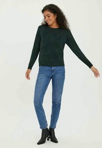 Suéter verde escuro texturizado com mangas longas, combinado com jeans ajustados azuis e botins pretos. O corte é casual e descontraído.