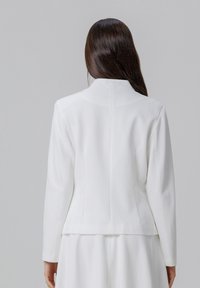 Blazer blanc avec une silhouette structurée, caractérisé par un col arrondi, des manches longues et un tissu lisse. Détails de coutures subtils à l'arrière.