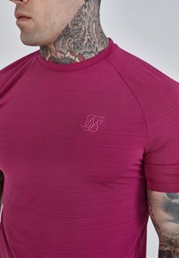 Pieguļošs īsās piedurknes krekls izstrādāts no teksturēta dziļi rozā auduma ar rievojuma rakstu. Arī uz krūtīm ir nemanāmi ievietots logo.