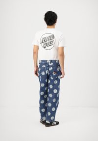 White t-shirt with black "Santa Cruz" logo, paired with blue jeans featuring white dice pattern; casual footwear with three black stripes.
