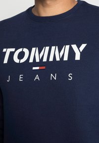 Marinblus långärmad skjorta som har fet vit text "TOMMY" med en röd och vit accent under, vilket indikerar varumärke, avslappnad stil och material.