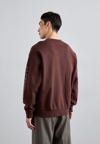 Καφέ φούτερ με άνετη εφαρμογή, ριμπ μανσέτες και το κόψιμο στο κάτω μέρος. Διαθέτει γραφική εκτύπωση κειμένου κατά μήκος του αριστερού μανικιού. Φοριέται με ανοιχτόχρωμα παν trousers.