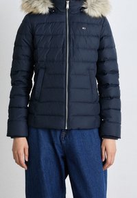 Navy puffer jacket with faux fur hood, zip closure, and vertical quilting. Paired with blue denim jeans.