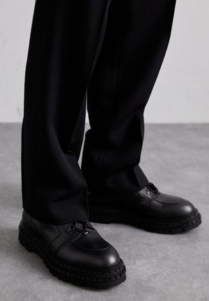 Pantofi din piele neagră, cu vârf rotund, suprafață texturată, design cu șireturi și talpă groasă din cauciuc cu un model de profil. Purtați cu pantaloni negri.