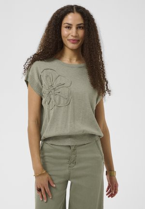 Kvinde med krøllet hår iført en lysegrøn striktop med stort blomsterdetalje og matchende grønne bukser, guldt armbånd og ringe.