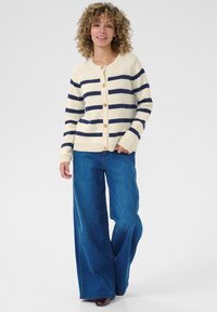 Gilet crème avec rayures bleues, col rond et boutons. Associé à un jean bleu à jambes larges. Texture en tricot douce.