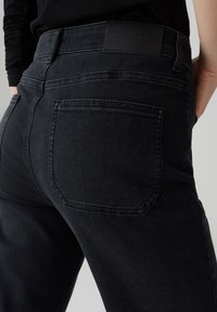 Close-up van een persoon die zwarte denim jeans draagt, waarbij de achterzak en een deel van de tailleband en heupgebied zichtbaar zijn.
