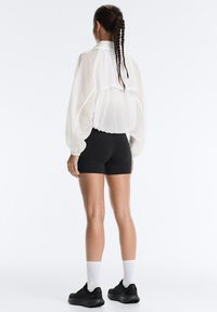 Femme aux cheveux tressés, vue de dos, portant une veste blanche transparente, un short noir, des chaussettes blanches et des chaussures de sport noires sur fond blanc.