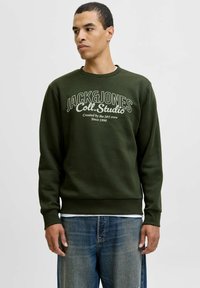 Mand iført en mørkegrøn crewneck sweatshirt med teksten "JACK & JONES Coll. Studio", parret med blå jeans, der står mod en lys baggrund.