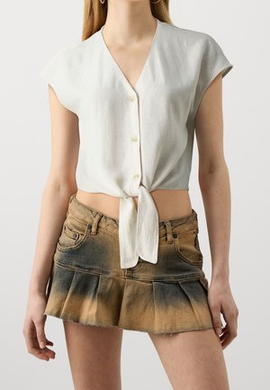 Femme portant un crop top blanc à manches courtes boutonné, noué à la taille, avec une mini-jupe plissée ombrée marron et noire.