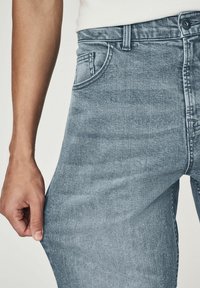 Jeans din denim de un albastru deschis, cu o textură ușor decolorată, având un design clasic cu cinci buzunare și un nasture vizibil la talie, ținuți de o mână.