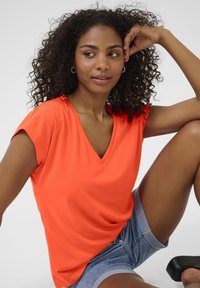 Bright orange, short-sleeved V-neck top made of smooth fabric, featuring gentle ruffles along the shoulders, paired with denim shorts.