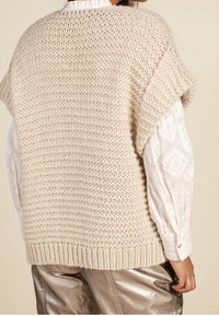 Pull beige tricoté oversize à manches courtes, avec un motif texturé et un ourlet côtelé. Porté sur un chemisier blanc à motifs.
