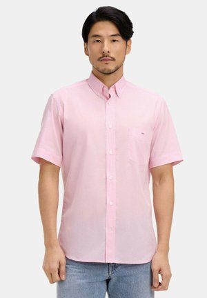 Homme portant une chemise rose clair à manches courtes avec une petite poche poitrine, associée à un jean bleu clair, debout devant un fond uni.