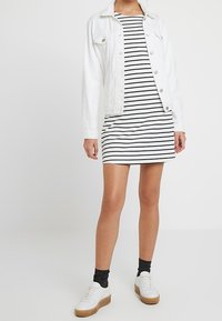 Valkoinen farkkutakki mustavalkoisen raidallisen mekon päällä. Mekko on lyhyt, ja asukokonaisuuteen on yhdistetty valkoiset tennarit ja mustat sukat.