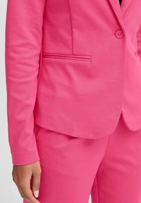 Světlý růžový blazer s jedním knoflíkem, přiléhavý střih a boční kapsou. Měkká textura s minimem designových ozdob.