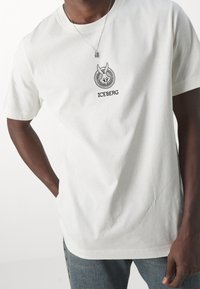 Biała bawełniana koszulka z okrągłym dekoltem, krótkimi rękawami oraz czarnym haftowanym logo na piersi z napisem „ICEBERG”.