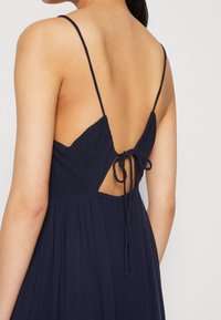 Marineblaues Rückenauschnittkleid mit dünnen Trägern, ausgestattet mit einer gebundenen Verschlusslösung am Rücken und weichem, fließendem Stoff, der einen plissierten Effekt erzeugt.