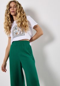 Witte cropped t-shirt met een bloemen- en bladprint. Groene wijdvallende broek van textuurstof met elastische tailleband.