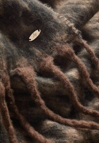 Écharpe en laine marron douce et duveteuse avec des franges et une petite étiquette ovale en métal attachée près d'un bord.